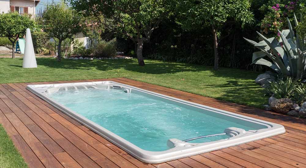 Un spa de nage est installé dans un jardin verdoyant. Il est entouré par une terrasse en bois.