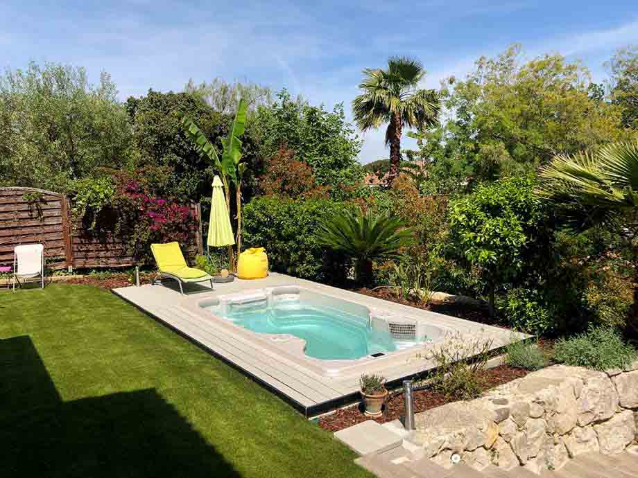 Un spa de nage est implanté dans un jardin verdoyant. Il est entouré par une terrasse claire. Une chaise longue et un parasol sont installés à proximité du spa de nage. 