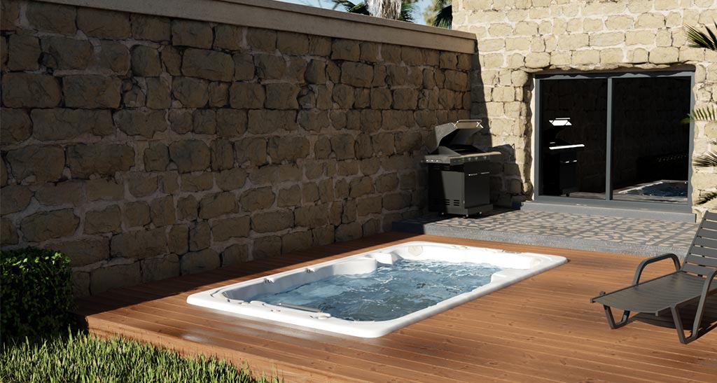 Spa de nage 13SE encastré dans une terrasse en bois dans le jardin d'une maison en pierres à côté d'un transat et d'un barbecue.