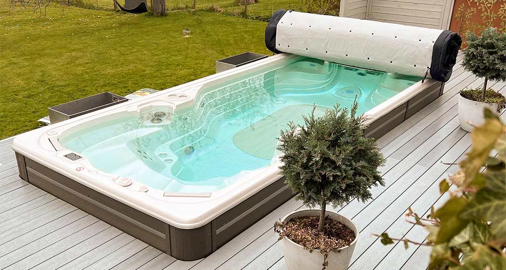 Spa de nage 17AX avec sa couverture enroulée encastré dans une terrasse dans un jardin avec deux pots de fleurs sur le côté.