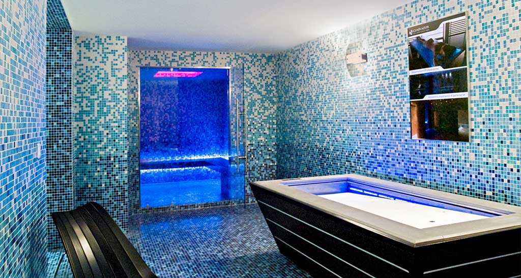 Hammam Ovation avec place assises et une lumière d'ambiance bleu avec une baignoire et un banc en premier plan.