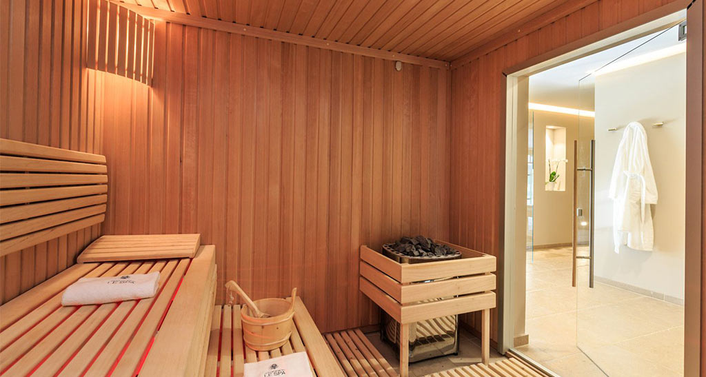 Sauna Lambris Sensation vu de l'intérieur avec un seau et un four. Une porte vitrée ouverte donnant sur un couloir avec un peignoir sur un support.