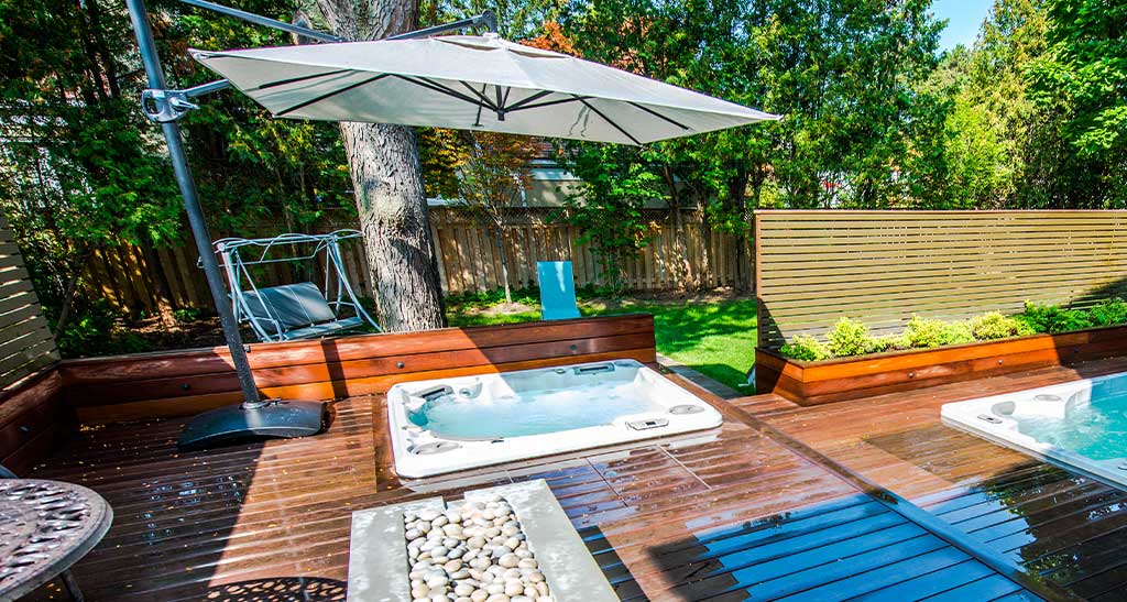 Deux spas encastrés dans une terrasse en bois avec un parasol au dessus et un bout de jardin en arrière-plan.