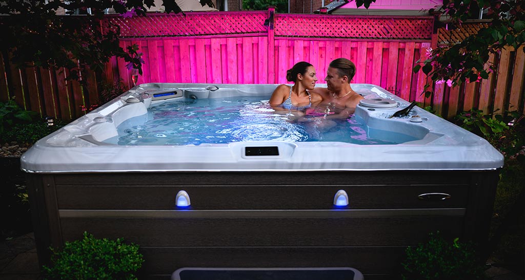 Couple se faisant un câlin dans un spa H720 en début de soirée avec une lumière tamisée rose qui donne sur un brise-vue en bois.