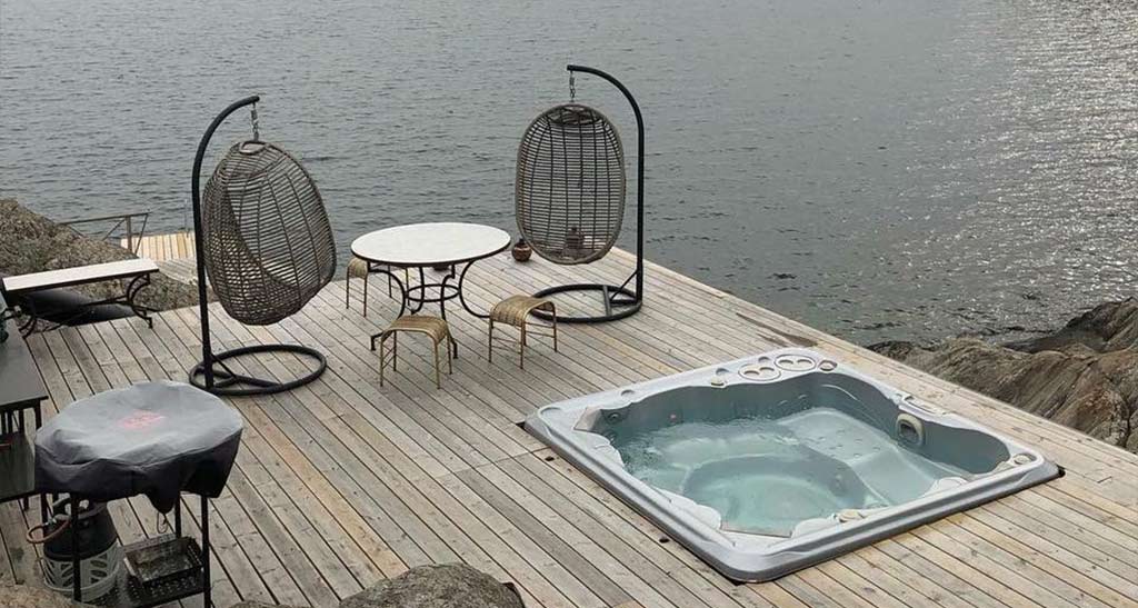 Spa H770 encastré dans une terrasse en bois aménagée d'une table avec ses tabourets et deux fauteuils œuf donnant sur la mer.