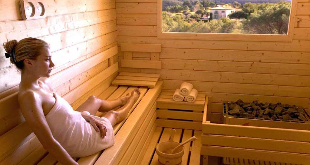 Femme en serviette assise dans un sauna Massif Prestige à côté d'un petit seau, d'un four et trois serviettes roulées et une vue sur une maison entourée de verdure.