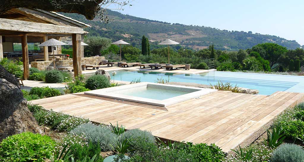 Spa mosaïque Plazza encastré dans une terrasse en bois avec une piscine à débordement en arrière-plan qui donne sur une montagne.