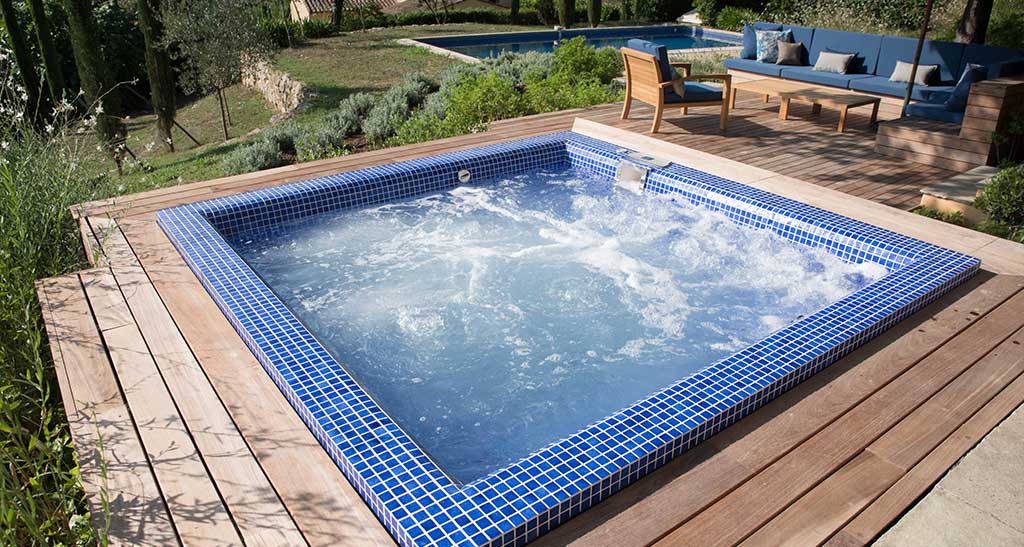 Spa mosaïque Plazza Lounge encastré dans une terrasse en bois à côté d'un salon de jardin, avec une piscine creusée en arrière-plan.