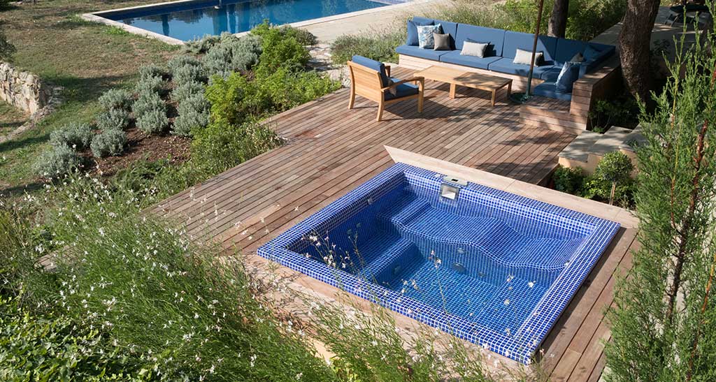 Spa mosaïque Plazza Lounge vu de haut encastré dans une terrasse en bois à côté d'un salon de jardin, avec une piscine creusée en arrière-plan.