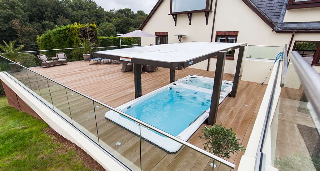 Abri Hydrocover pour spa de nage encastré ouvert sur une terrasse en bois à côté d'une maison et de la verdure en arrière plan
