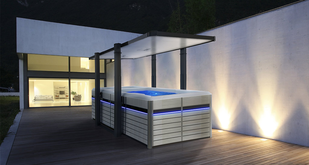 Abri Hydrocover pour spa de nage ouvert sur une terrasse en bois avec une baie vitrée de maison en arrière plan la nuit