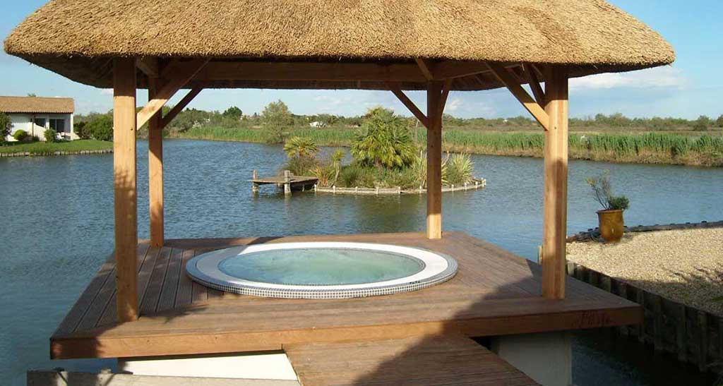 Spa à débordement Mosaic 250 encastré dans une terrasse en bois sous une pergola à côté d'un lac.