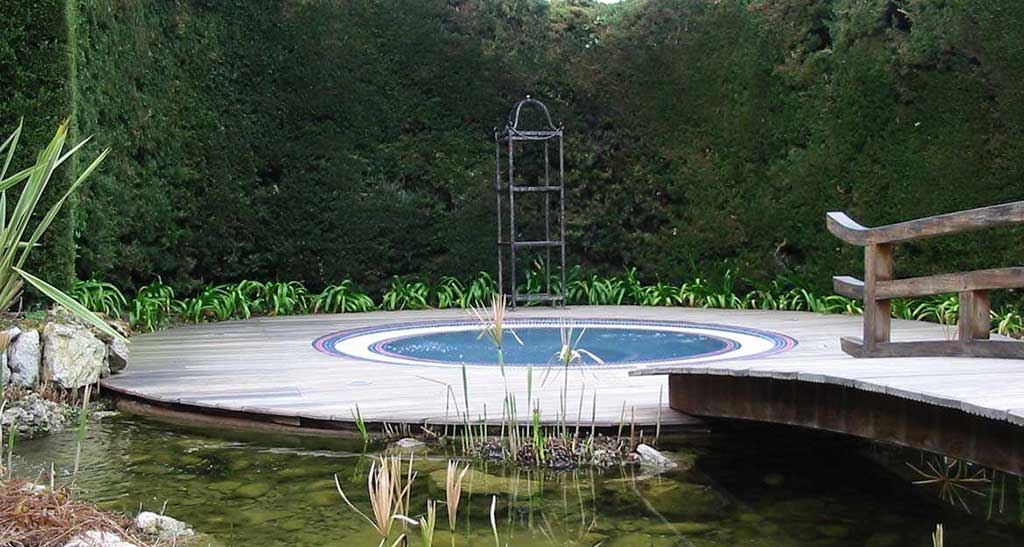 Spa à débordement Mosaic 250 encastré dans un coin de jardin à côté d'un étang et de verdure.