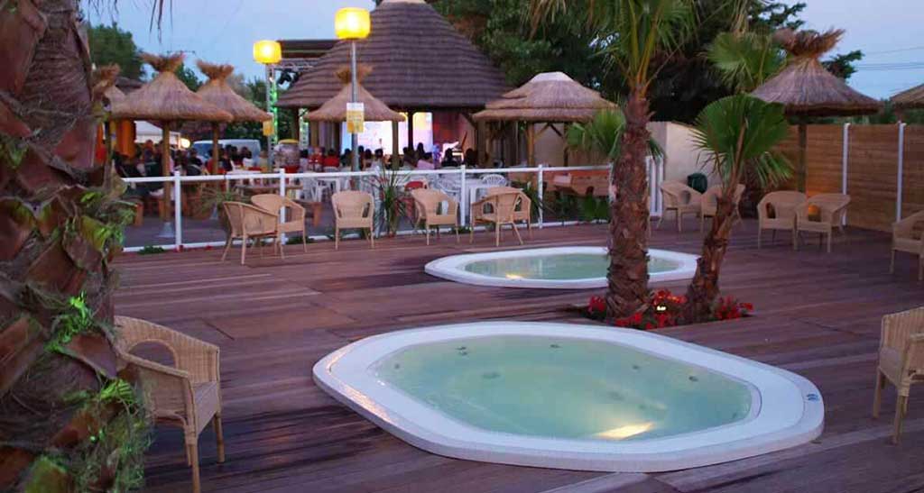 Spa à débordement Mosaic 350 encastré dans une terrasse en bois à côté de palmiers et chaises d'extérieur et un restaurant en arrière plan.