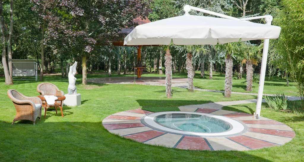 Spa à débordement Mosaic 360 encastré dans un jardin plein de verdure sous un parasol avec deux chaises d'extérieur à côté.