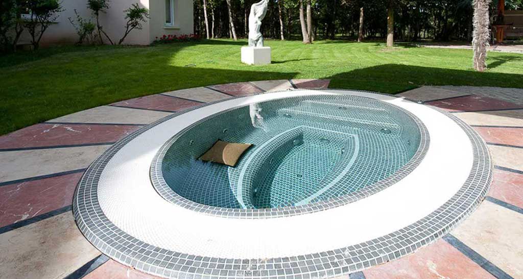 Spa à débordement Mosaic 360 encastré dans un patio dans un jardin avec une statue en arrière plan et un coussin dans l'eau.