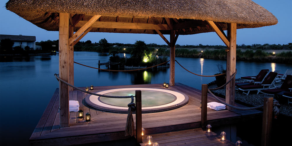 Spa mosaïque encastré dans une terrasse en bois avec une tonnelle au-dessus et un étang à côté, le tout de nuit avec des bougies autour