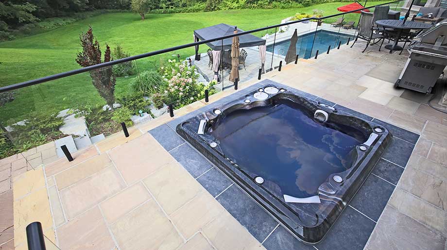 Un spa est encastré dans une terrasse, à proximité d'un salon de jardin et d'un barbecue. Il offre une vue sur le jardin, la pergola et la piscine installée plus bas. 