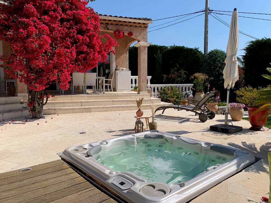 Un spa est installé sur une terrasse. Le bassin est encastré. Il est situé à proximité d'une maison et d'un bougainvillier rose en fleurs. 