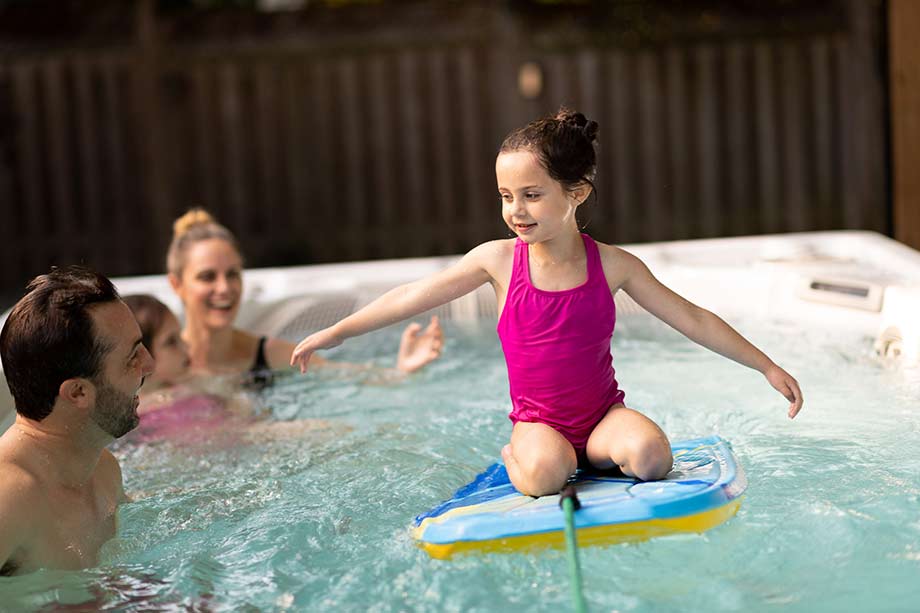 Une enfant et sa famille sont dans un spa de nage. La petite fille est agenouillée sur un bodyboard et tente de garder l'équilibre. 