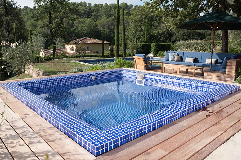 Quelle est la consommation d'eau d'un spa ? Un spa recouvert de mosaïque bleu marine est encastré dans une terrasse en bois clair. Il est placé dans un jardin, à proximité d'un salon de jardin. 