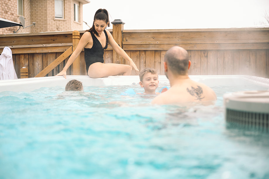 Une famille se baigne dans un spa de nage placé en extérieur. On peut voir de la vapeur s'en dégager. 