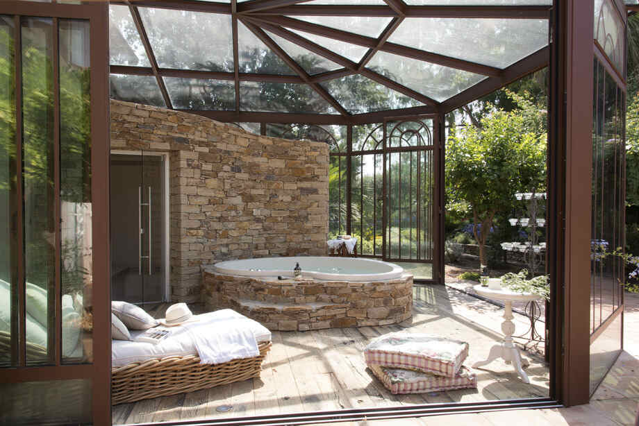 Un spa intérieur avec un habillage en pierre naturelle est installé sous une véranda en verre et en acier. 