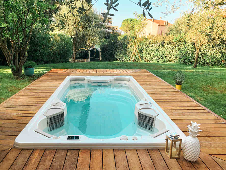 Un spa de nage est placé dans un jardin. Il est entouré par une terrasse en bois. 