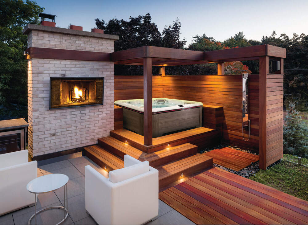 Un spa est installé en extérieur sur une terrasse surélevée, à proximité d'une cheminée et d'un salon de jardin. 