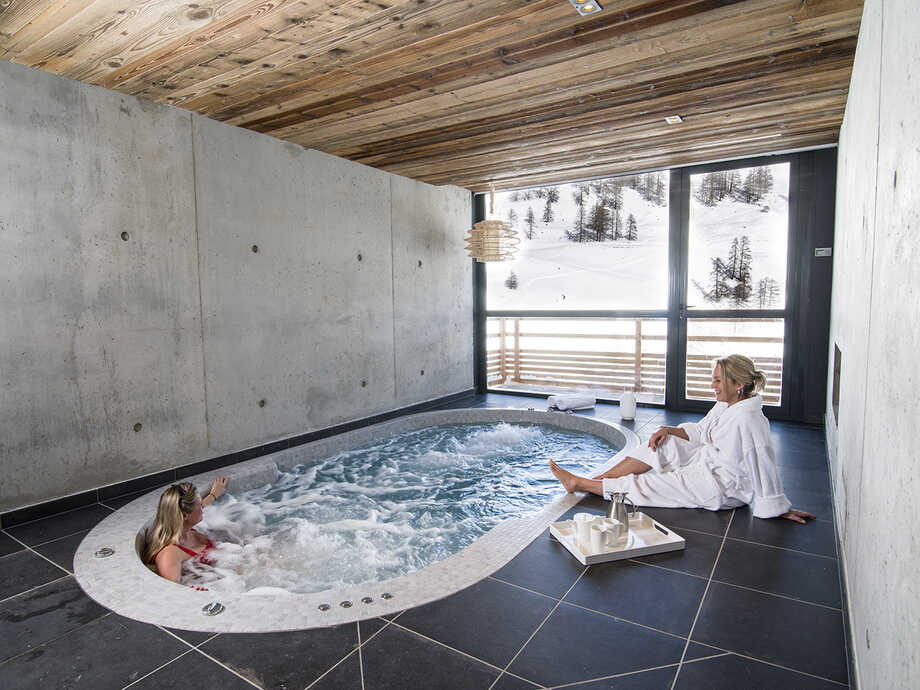 Un spa de nage est installé en intérieur face à une baie vitrée qui offre une vue sur les montagnes enneigées. 