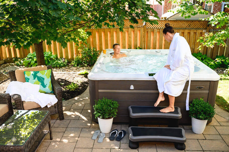 Un spa hors sol est installé dans un jardin. Un homme se détend dans le spa tandis qu'une femme habillée d'un peignoir utilise la platine du spa pour faire des réglagles. 